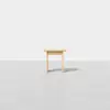 A small, simple wooden stool with three legs stands on a smooth, light gray floor against a plain white background.