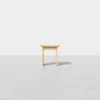 A small, simple wooden stool with three legs stands on a smooth, light gray floor against a plain white background.