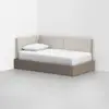 A daybed with a light beige upholstered backrest and side, single white pillow, and white bedding, set in the corner of a minimal, white-walled room with a light gray floor.