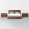 A minimalist bedroom setup featuring a wooden bed frame with white bedding and two matching wooden nightstands on either side, all set against a plain white wall and floor.