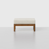 A modern ottoman with a light beige cushioned top and a simple wooden base, set against a plain white background.
