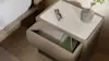 A bedside table with an open drawer, a green book inside, and a white tabletop holding black eyeglasses and a small gray vase with a leafy branch. The edge of a bed is visible on the left.