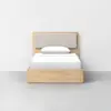 A minimalist wooden bed with a light fabric headboard, white bedding, and a single pillow, positioned against a plain white wall on a gray floor.