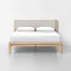 A minimalist wooden bed frame with a light beige upholstered headboard, neatly made with white bedding and two white pillows, set against a plain white wall and floor.