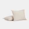 Two square, light beige pillows with a soft fabric texture are placed on a plain white background; one pillow is standing upright, and the other is lying flat in front of it.