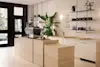 Minimalist coffee shop interior with light wood counters, white cabinetry, a large espresso machine, potted plants, shelves with coffee bags, and a menu on the wall. Natural light streams in through large windows.