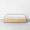 A minimalist bed with a light wooden frame, white mattress, and two white pillows is set against a plain white wall on a light gray floor.