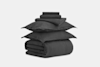 A neatly stacked set of dark gray bedding, including two pillows, a folded duvet, a flat sheet, a fitted sheet, and two pillowcases, arranged against a plain white background.