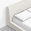 Close-up view of a neatly made bed with white sheets and pillows, featuring a light beige upholstered headboard and frame, placed on a light-colored floor against a white wall.