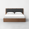 A modern wooden bed with a simple frame, a dark upholstered headboard, white sheets, and two white pillows, set against a plain white wall and grey floor.