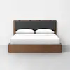 A modern wooden bed with a simple frame, a dark upholstered headboard, white sheets, and two white pillows, set against a plain white wall and grey floor.