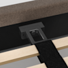 Close-up of a bed frame showing a black metal support bracket attached to wooden slats and the upholstered bed base, with a black strap laid across the slats.