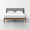 A modern wooden bed frame with a gray upholstered headboard, neatly made with white bedding and two white pillows, placed in a minimalistic room with a white wall and floor.