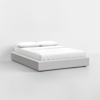 A modern, low-profile bed with a light gray upholstered frame, dressed in white bedding and two white pillows, set against a plain white background.