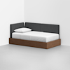 A modern daybed is placed in a corner, featuring a wooden base, white mattress and bedding, a single pillow, and dark gray upholstered back and side panels against a plain white wall and floor.