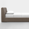 A side view of a modern, minimalist bed with a gray upholstered frame, a white mattress, white sheets, and a white pillow, set against a plain white wall and a light gray floor.