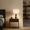 A modern bedroom with a wooden nightstand beside a bed, featuring a decorative stone-based table lamp with a white shade, a candle, and a small tray on the nightstand. Shelves are visible in the background.