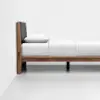 A modern wooden bed frame with a dark upholstered headboard, white bedding, and a single pillow, positioned against a white wall on a light floor.