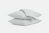 Two light gray pillows are stacked on top of each other, with a folded duvet or blanket draped over the top pillow, all set against a plain white background.