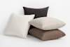 Four textured square pillows are stacked and leaning against each other on a white background. The pillows are in shades of cream, beige, brown, and dark brown.
