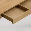 A close-up view of a wooden desk with an open drawer, showing an empty interior with dividers. The desk and drawer are made of light-colored wood, set against a plain, light background.