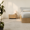 A minimalist bedroom features a wooden bed frame and matching nightstand with a modern table lamp, a mug, and a book. Soft sunlight streams in, highlighting neutral tones and a leafy plant in the corner.