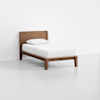 A minimalist wooden bed frame with a headboard, dressed with a white mattress, white sheets, and a white pillow, set in a plain, light-colored room.