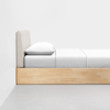 A minimalist wooden bed with a beige upholstered headboard, neatly made with white bedding and a single pillow, positioned against a white wall on a gray floor.