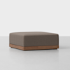 A square, brown upholstered ottoman with a wooden base sits on a light gray floor against a plain white background.