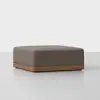 A square, brown upholstered ottoman with a wooden base sits on a light gray floor against a plain white background.