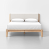 A minimalist bed with a light wooden frame and a beige upholstered headboard, neatly made with white bedding and two pillows, set against a plain white wall and floor.