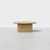 A minimalist wooden table with a round top and a solid rectangular base, placed on a light gray floor against a plain white background.