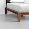 Close-up of a modern wooden bed frame with tapered legs, a white mattress, and white bedding in a minimalistic room with a light gray floor and a plain white wall.