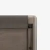 Close-up of the top corner of a modern wooden crib with a rounded edge and a gray fabric rail cover, set against a plain white background.
