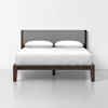 A modern wooden bed frame with a gray upholstered headboard, white bedding, and two white pillows, placed against a plain white wall in a minimalistic room with a light gray floor.