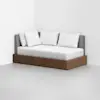 A modern daybed with a wooden base, gray upholstered backrest, and four white pillows, placed in the corner of a minimalist white room with light gray flooring.