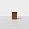 A minimalist wooden bench with a light-colored cushioned seat, positioned in the center of a plain, light gray room with a white background.