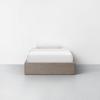 A single bed with a wooden frame, white bedding, and a white pillow, set against a plain white wall and gray floor in a minimalistic room.