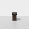A minimalist, dark wood bench with a black cushioned seat is centered against a plain, light gray background. The bench casts a faint shadow on the smooth floor.
