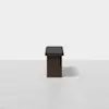 A minimalist, dark wood bench with a black cushioned seat is centered against a plain, light gray background. The bench casts a faint shadow on the smooth floor.