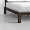 A close-up view of a modern wooden bed frame with dark brown legs, supporting a neatly made bed with white bedding in a minimalist, light-colored room.