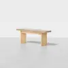 A minimalist wooden bench with a light wood finish and a flat, rectangular seat, set against a plain, light gray background.