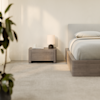 A modern bedroom with a light gray bed, matching wooden nightstand, a small table lamp, a book, and a cup. Soft natural light filters in through curtains, and a leafy plant sits in the foreground.