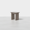 A small, modern side table with a rectangular top and three wide, flat legs, all in a smooth, light gray wood finish. The table is placed in a minimal, light-colored setting.