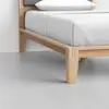 A close-up view of a wooden bed frame with a white mattress and pillow, set on a light grey floor in a minimalist bedroom.