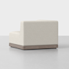 A modern armless corner sofa with light beige upholstery and a wooden base, displayed in a minimalistic, light gray setting.
