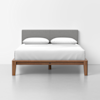 A modern bed with a light gray upholstered headboard, white bedding, two white pillows, and a wooden frame, set against a plain white wall and floor.