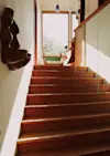 A sunlit wooden staircase leads up to a landing with a cushioned chair by a large window. Sculptural decor hangs on the left wall, and a glass railing lines the right side.