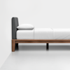 A minimalist bed with a dark gray upholstered headboard, white bedding, and a wooden frame on a light-colored floor against a plain white wall.