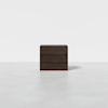 A minimalist dark wood three-drawer dresser is centered in an empty, bright room with a light gray floor and a plain white background.
